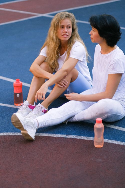 Free Full body of confident young female friends in activewear sitting and talking on sports ground after training with water bottles in daytime Stock Photo