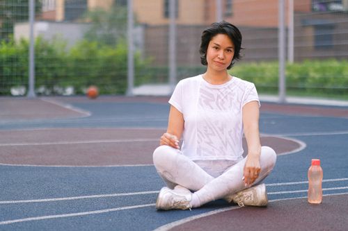 Free Full body of young female in sportswear with water bottle sitting with crossed legs on sports ground near fence and looking at camera in daytime Stock Photo