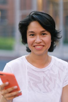 Happy young woman in casual outfit text messaging on smartphone in daytime outside and looking at camera
