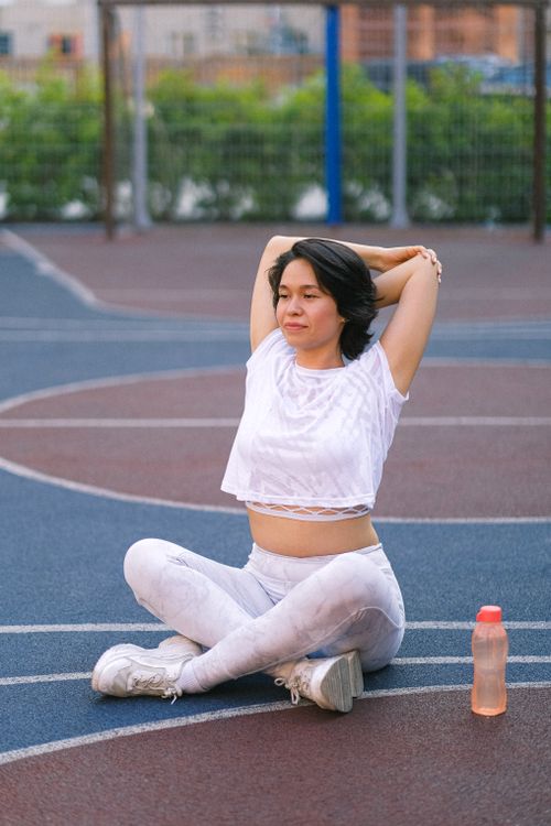 Free Full length of young woman in sportswear training on court near bottle with water with crossed legs in daylight near fence Stock Photo