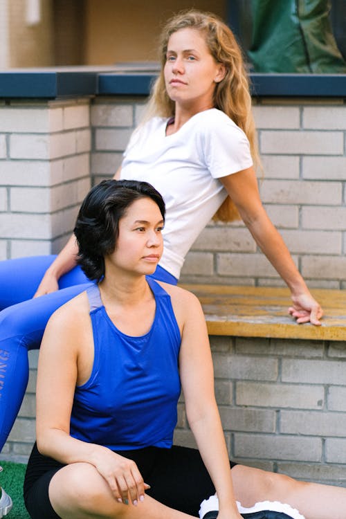 Attractive young sportswomen in activewear resting together on bench and grassy ground in park after training
