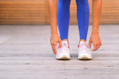 Crop anonymous sportswoman in blue leggings and sneakers stretching body and bending forward while warming up before exercising in fitness studio