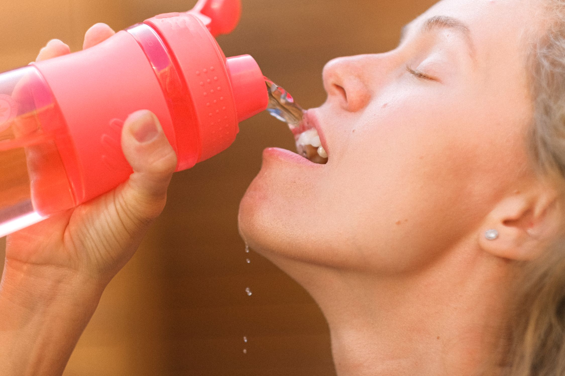 Tired young woman pouring water into mouth from reusable bottle after outdoor training