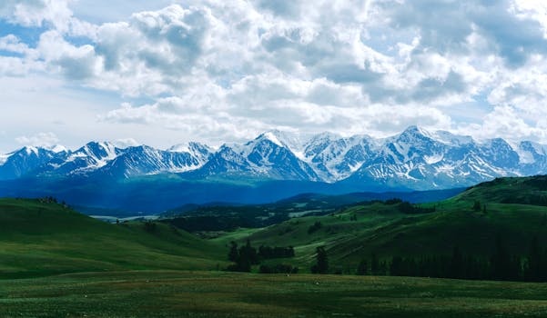 Stunning snow-capped mountain range surrounded by lush green valleys under cloudy skies.