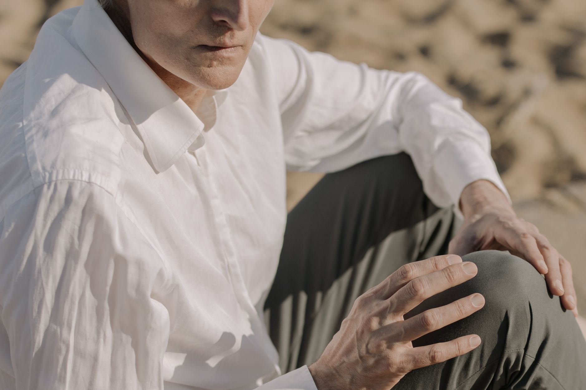 Close-up of an unrecognizable man sitting on a sandy beach in white shirt and gray pants.