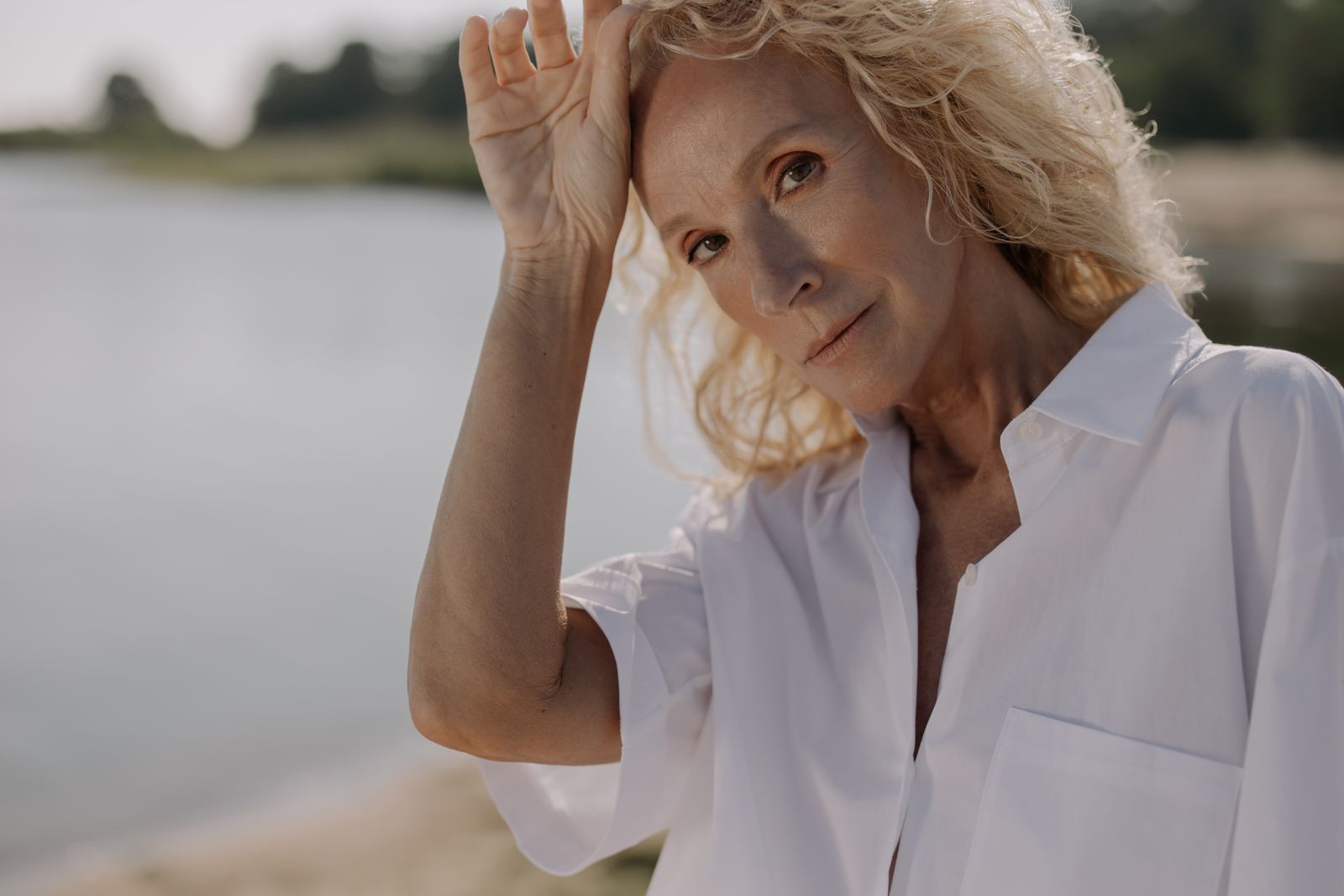 Woman with curly blonde hair in a white shirt resting her hand on her head beside a lake in sunlight
