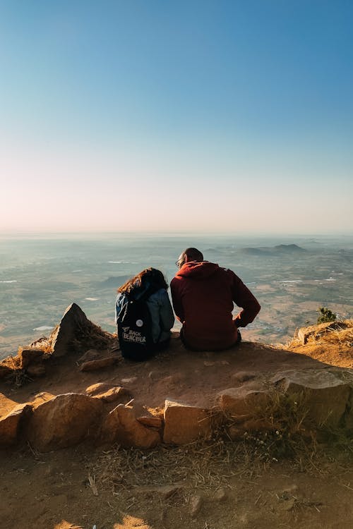 Back view of unrecognizable travelers in casual clothes sitting on stony coast near frozen  transparent lake against blue sky