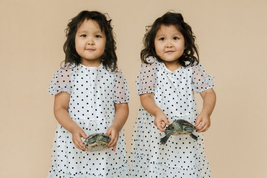 Adorable twin girls in polka dot dresses happily holding turtles in a studio setting.