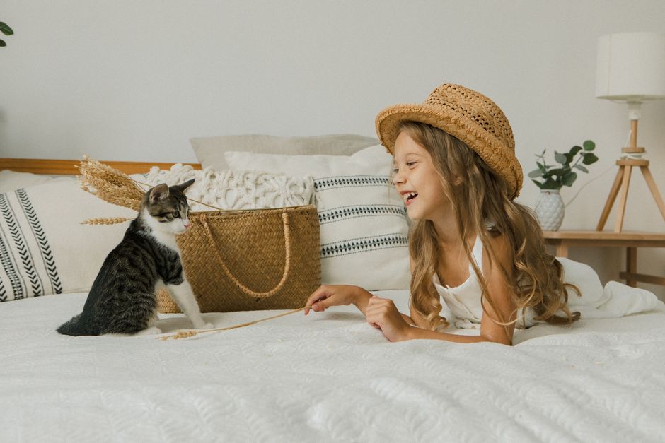 Żwirek sklep zoologiczny – gdzie kupić najlepszy w Polsce 3 Joyful child in a straw hat playing with a cat on a bed, surrounded by cozy decor and plants.