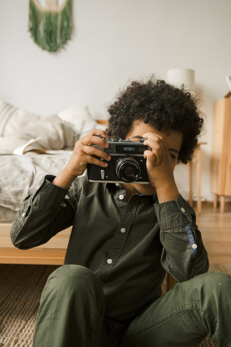 Little Boy Sitting In A Bedroom And Taking A Picture With A Camera 