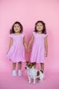 一对年轻的双胞胎女孩穿着相配的粉色连衣裙，手牵手站在粉色背景前，身旁有一只杰克拉塞尔梗犬