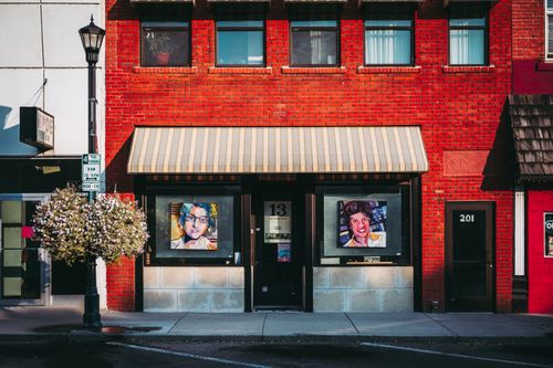 Free Exterior of red brick building with commercial space on ground floor with poster on door and pictures on windows located next to road on sunny day Stock Photo
