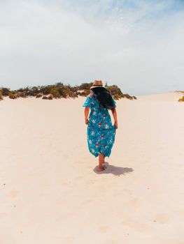A woman in a floral dress and hat walks across sandy dunes under a clear sky.
