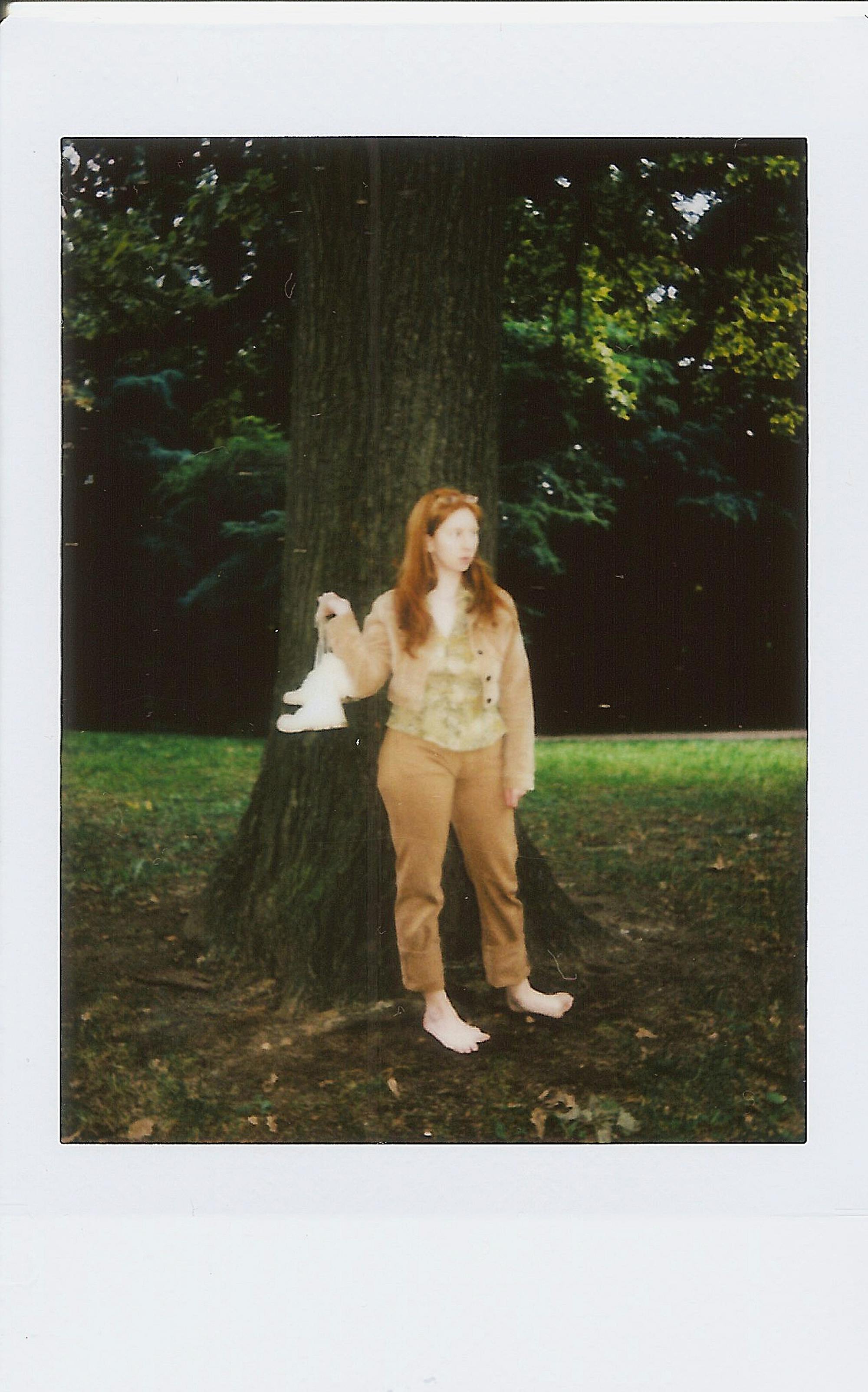 A woman holding a toy, standing barefoot by a tree in a forest setting.