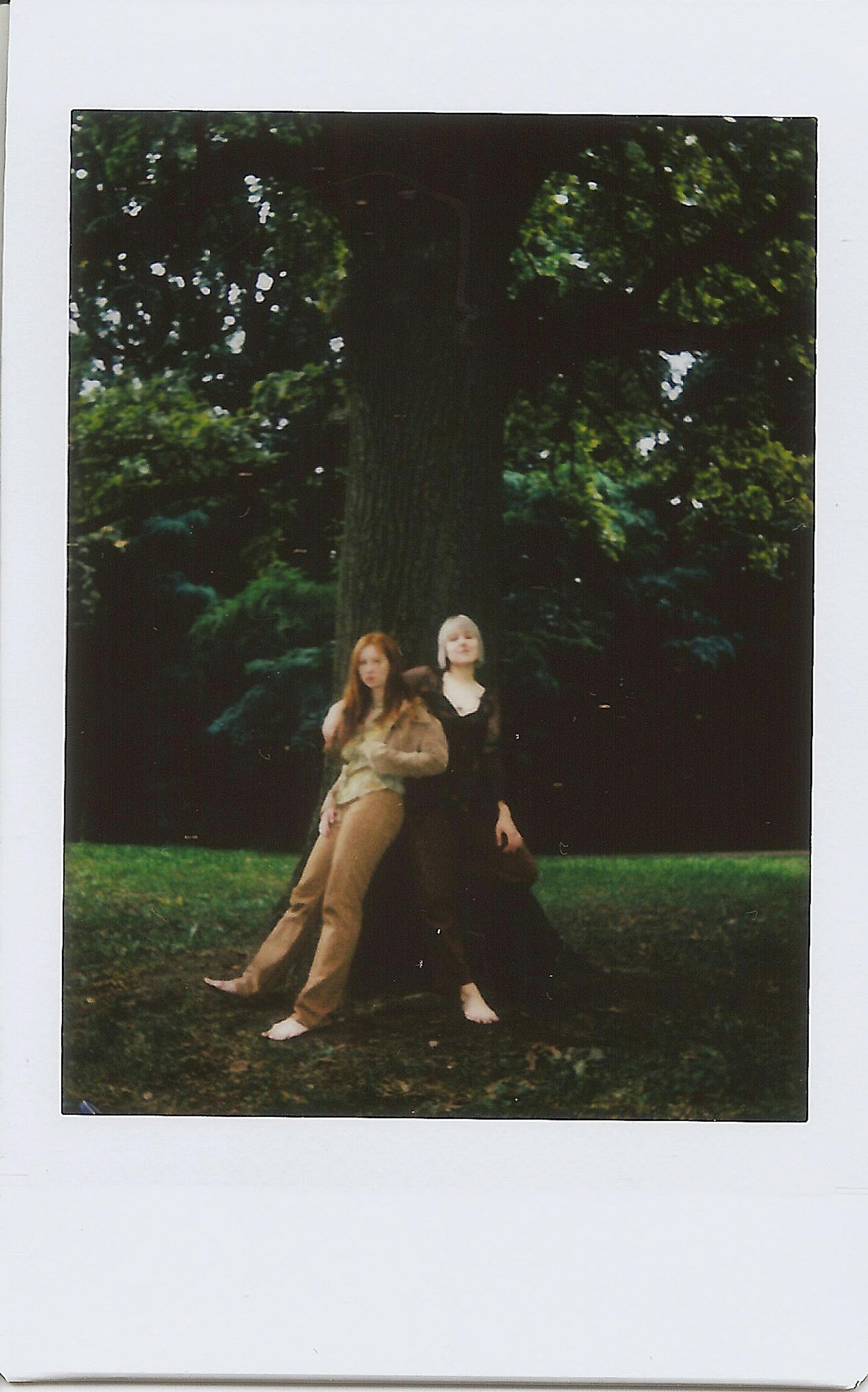 Two women in fashionable attire pose by a large tree, surrounded by lush greenery.