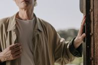 阳光下，一位穿着夹克的老人双手可见地站在户外，镜头特写