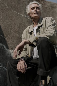 Portrait of a stylish senior man in a trench coat sitting against a stone backdrop. Outdoors setting.