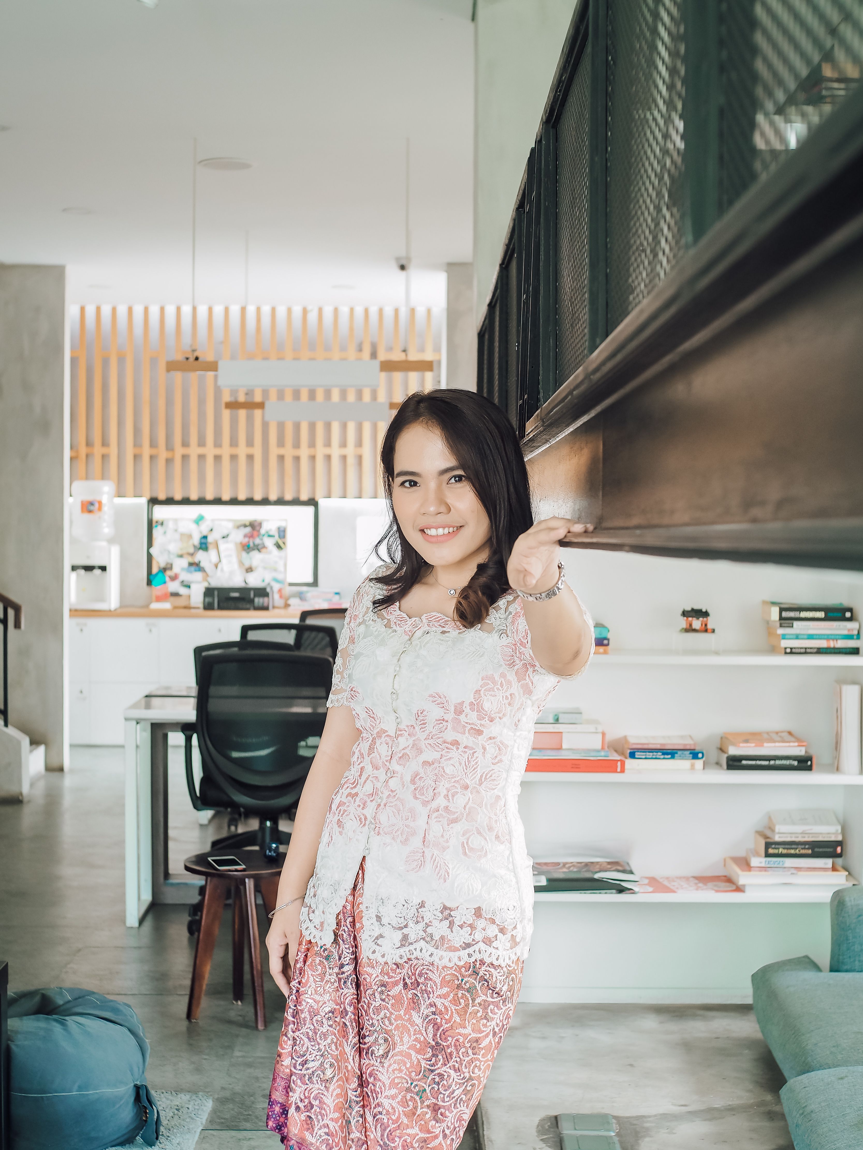 Asian woman smiling indoors, wearing an elegant white top with a lace design, in a modern home setting.