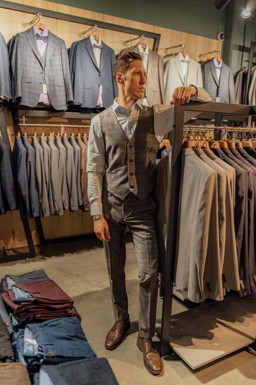 Free A fashionable man in a clothing store is picking a tailored suit from a rack, surrounded by various elegant options. Stock Photo