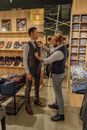 在一家时尚的男装精品店里，一名裁缝正在为顾客调整领带，四周摆满了衬衫和鞋子