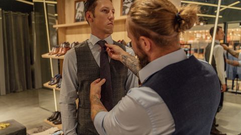 A tailor assists a client by adjusting his necktie in a clothing store fitting session.