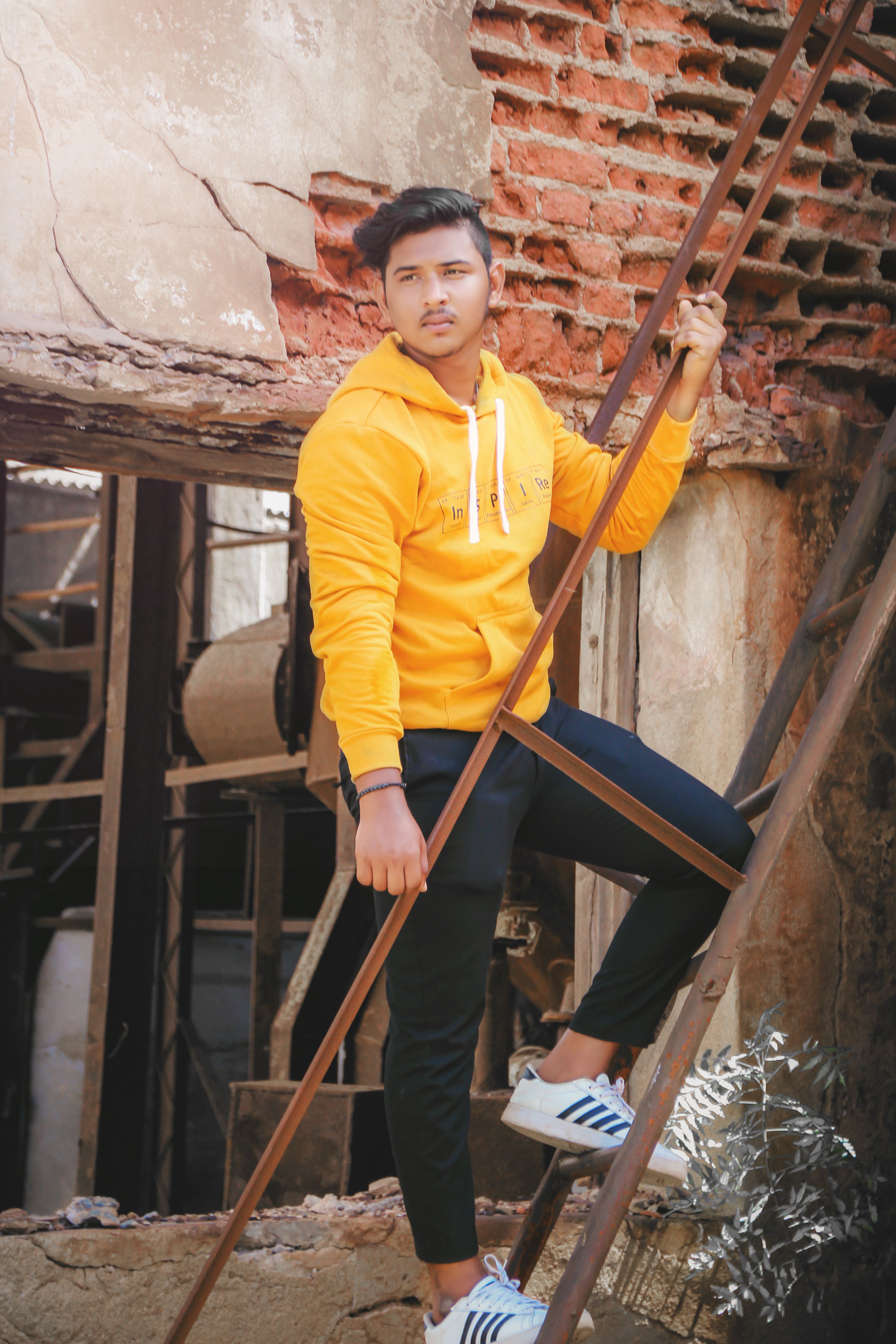 Man in yellow sweatshirt stands confidently on ladder against rustic urban backdrop.