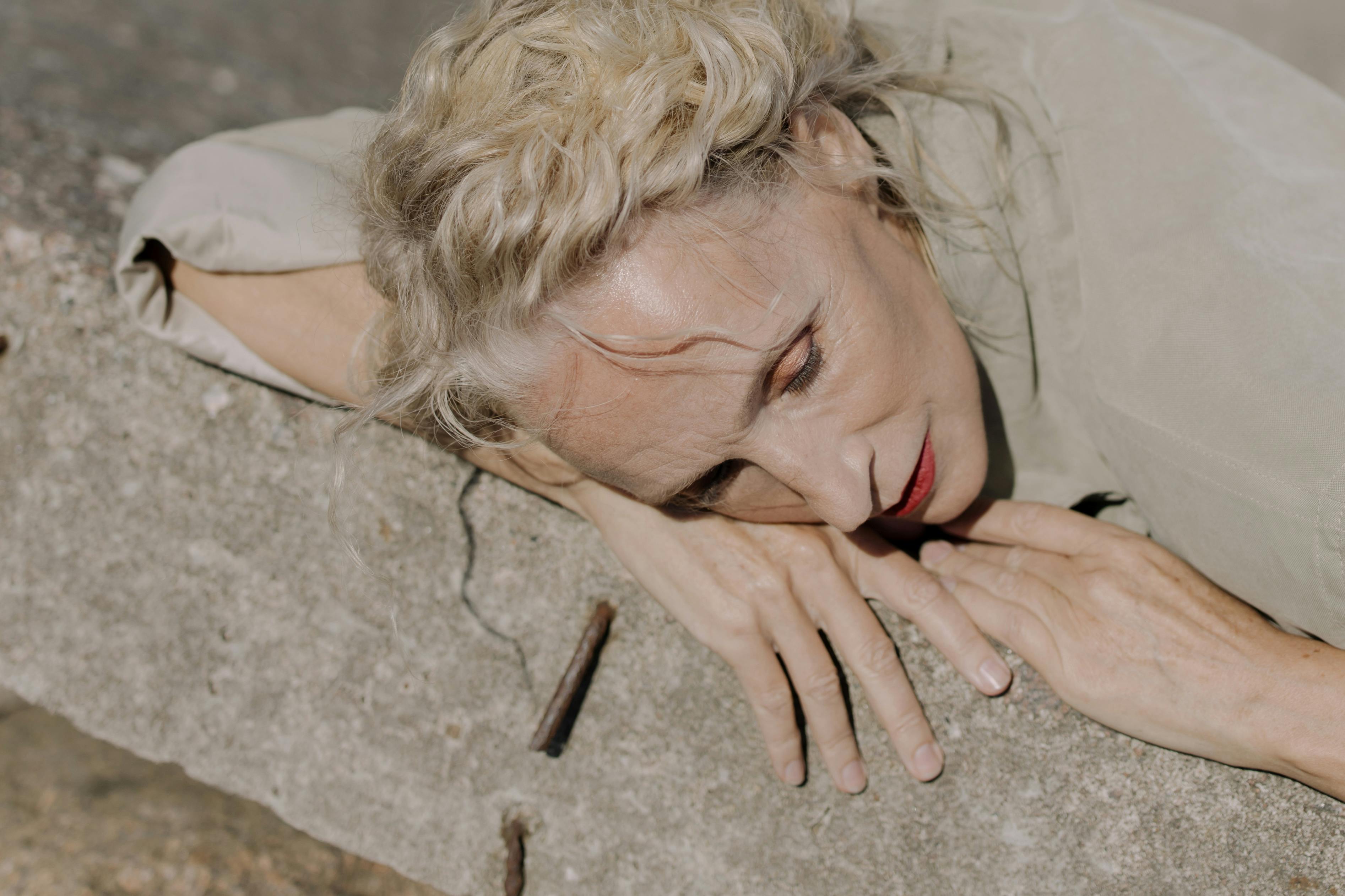 A graceful mature woman with gray hair rests her head on a concrete wall in an outdoor setting.