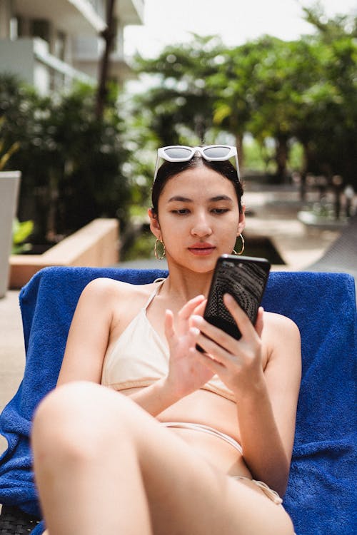 Graceful young ethnic female tourist in bikini messaging on smartphone while relaxing on deckchair on hotel terrace in sunlight