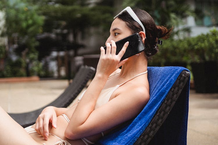 Young Slim Ethnic Lady Talking On Mobile Phone While Sitting On Sunbed In Hotel Yard