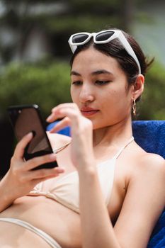 一位穿着比基尼的年轻少数族裔女游客在酒店院子的躺椅上放松，一边用手机看新闻