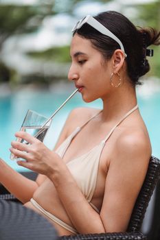 Side view of young sensual ethnic lady in stylish swimwear and sunglasses on head drinking cold water with straw while chilling near swimming pool during summer vacation