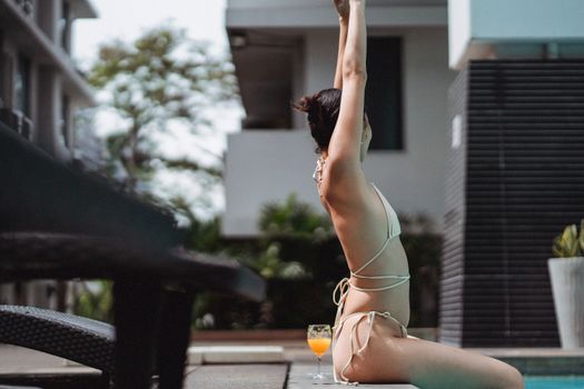 Side view of unrecognizable slim young female in bikini stretching arms while relaxing on edge of outdoor swimming pool during summer holidays in stylish resort hotel