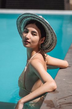 Side view of self assured young ethnic female traveler in stylish bikini and hat relaxing in swimming pool during summer holidays