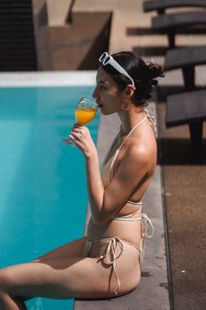 Side view of relaxed tanned young ethnic woman in bikini drinking fresh orange juice while sitting on pool border on sunny day