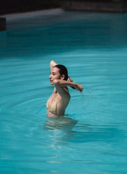 Side view of young graceful ethnic lady in bikini touching wet long hair while standing in swimming pool on sunny day