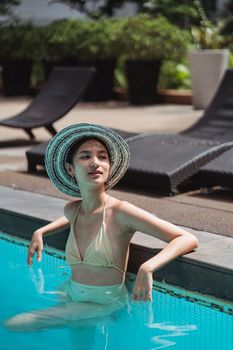 Slender young female in bikini and hat chilling in pool and leaning on poolside while looking away dreamily in sunny resort