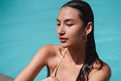 Beautiful young brunette with wet hair chilling in pool and leaning on poolside on hot sunny day