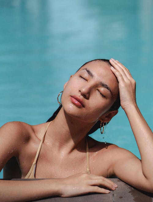 Relaxed woman leaning on poolside and touching head tenderly while chilling with eyes closed in pool and enjoying sunny day