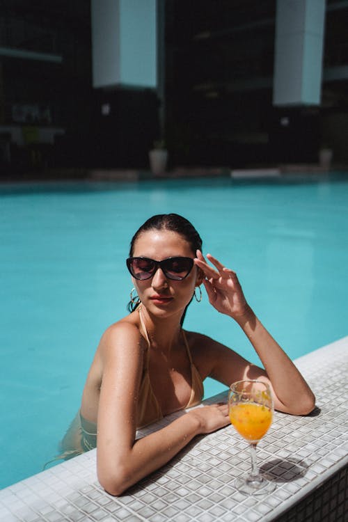 Free Beautiful tanned female wearing bikini and trendy sunglasses relaxing in outdoor pool with glass of refreshing orange juice while touching face gently Stock Photo
