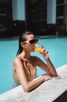 Side view attractive fit brunette in modish sunglasses chilling in outdoors swimming pool and sipping sweet orange juice on sunny day
