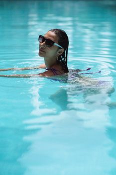 Side view young slim female in trendy sunglasses swimming in pool on clear day and looking at camera contentedly