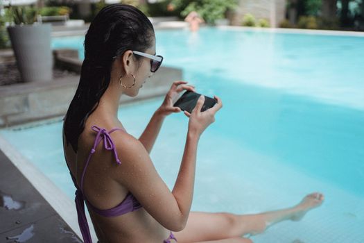 Side view attractive slim female wearing bikini relaxing on sunny resort poolside and taking photos of legs