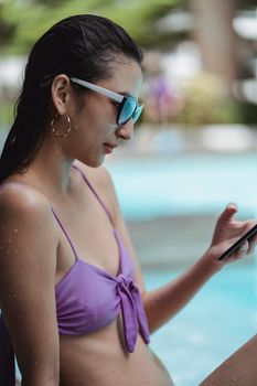 Side view of sensual young ethnic female with wet hair in trendy swimwear and sunglasses browsing mobile phone at poolside during summer holidays
