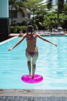 Back view of anonymous young fit lady in swimming wear jumping into pool water with rubber ring during summer holidays in resort