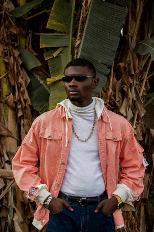 Serious young black male in trendy clothes and sunglasses standing with hands in pockets against palm trees in daytime