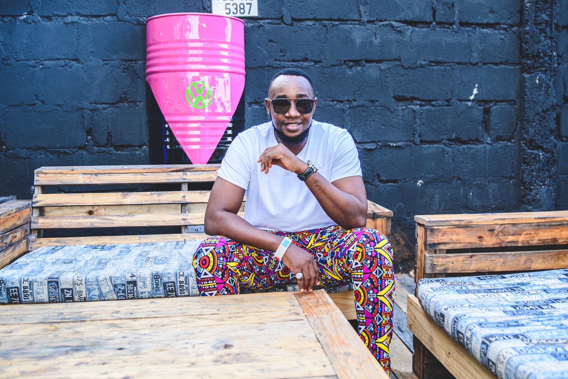 Smiling man wearing sunglasses and vibrant pants sitting on a wooden bench. Cool urban vibe in Dar es Salaam.