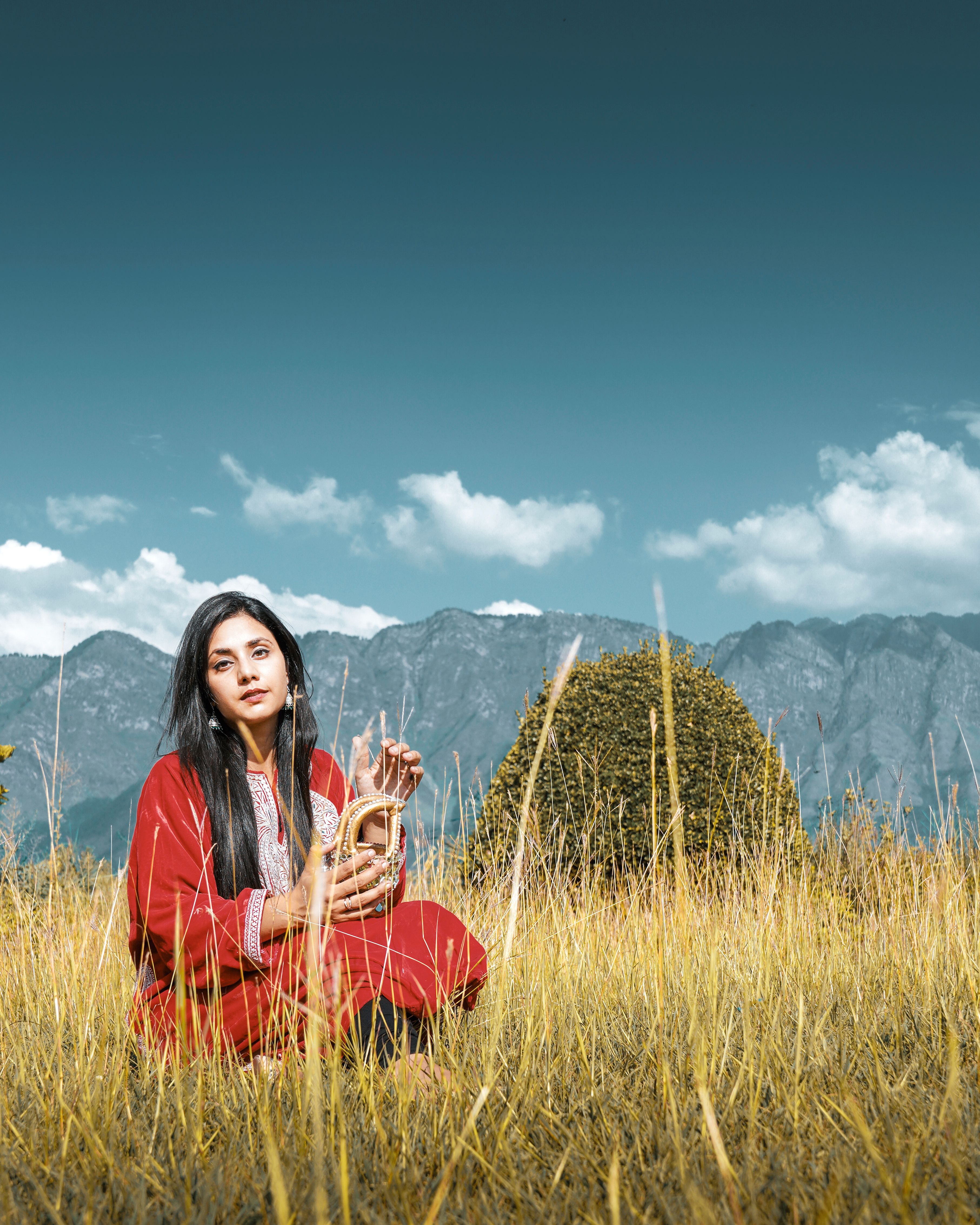 Woman in traditional attire sitting in a grassy field with mountains and sky backdrop.