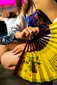 一名身份不明的女子穿着鲜艳的正品服装，赤脚蹲在背景模糊的咖啡馆里，手里拿着一把黄扇子