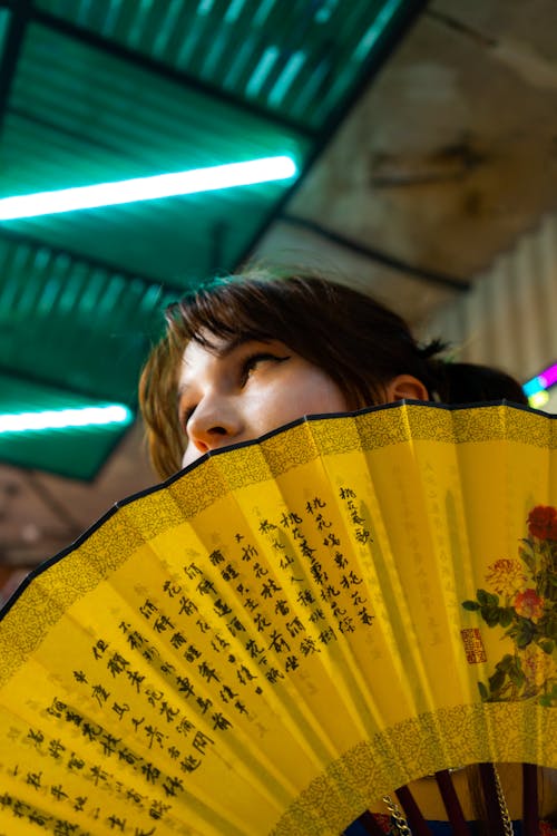 From below of young anonymous female with dark hair looking away while covering face with yellow fan with blurred ceiling