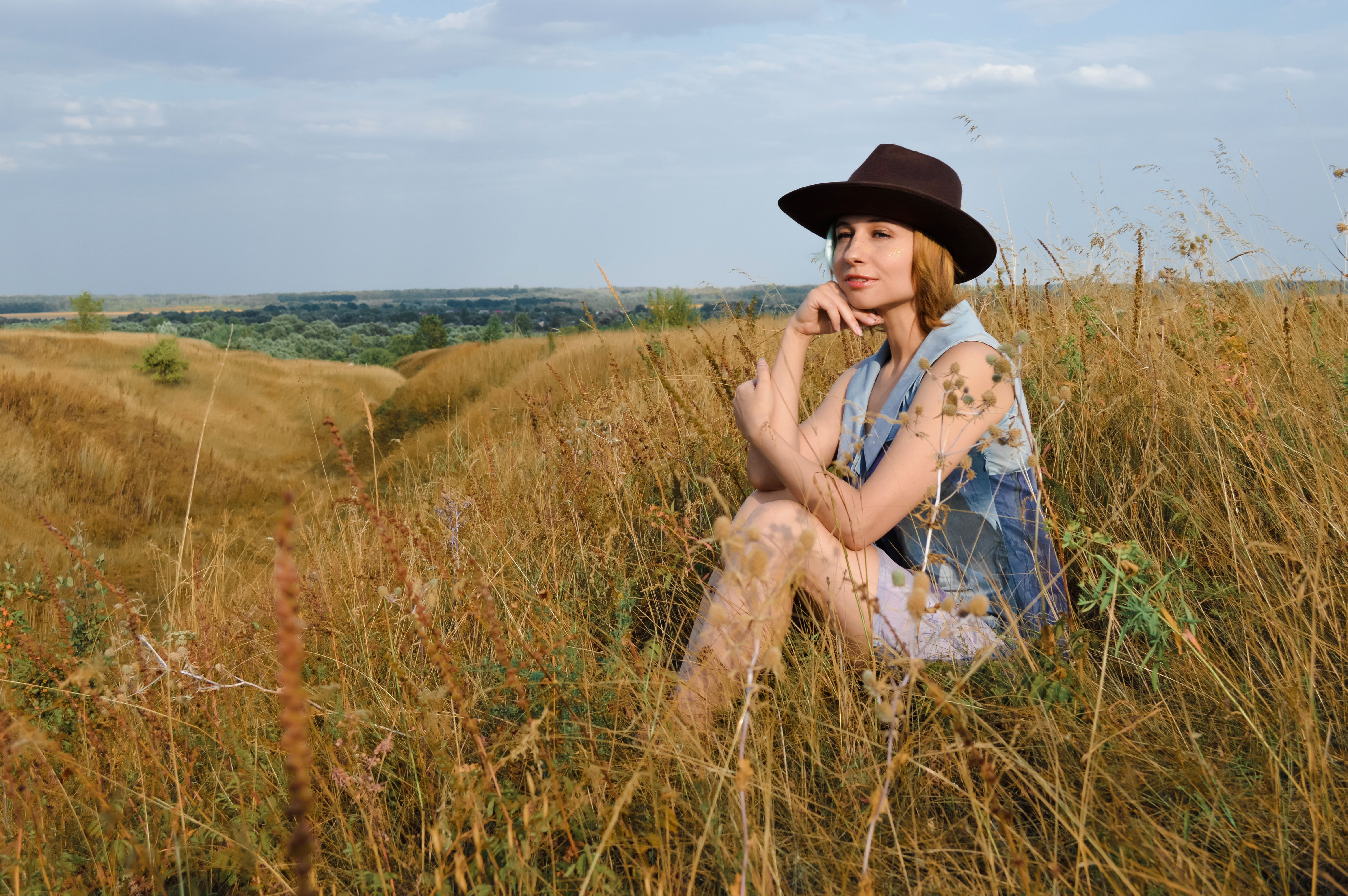 Full body stylish young female in brown hat and blue wear sitting on grass in meadow and looking away touching chin